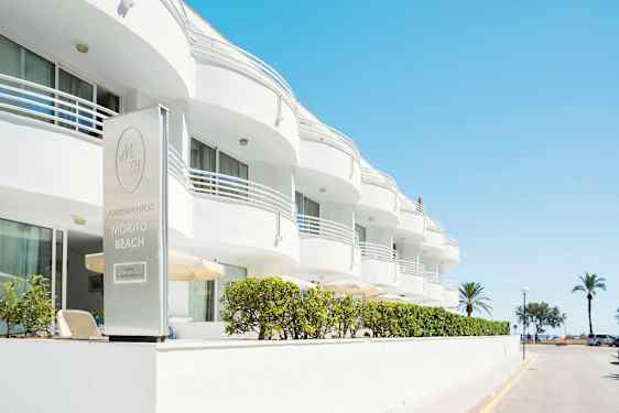 Den hvide bygning tilhører Morito Beach Apartments med palmer og hav i baggrunden. (AI-genereret tekst)