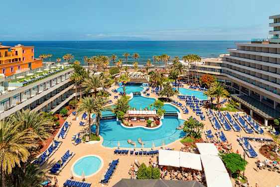 Det er udsigt over et hotelområde med flere swimmingpools, omkranset af liggestole og parasoller samt træer og bygninger. I baggrunden ses havet. (AI-genereret tekst)