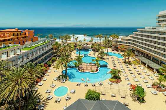 Et hotelkompleks med pools, liggestole og palmer, tæt på havet. (AI-genereret tekst)