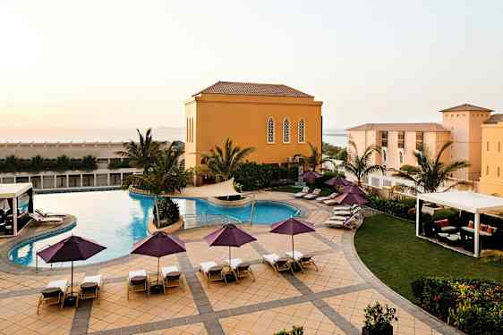 En udendørs pool med liggestole og parasoller er omgivet af palmer og en bygning med gule facader. (AI-genereret tekst)