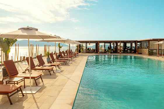 Billedet viser en udendørs pool med liggestole og parasoller langs poolkanten, med havet og en skyet himmel i baggrunden. (AI-genereret tekst)