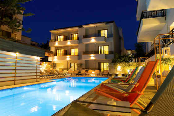 Billedet viser en oplyst hotelbygning og en udendørs pool omgivet af solsenge og loungestole om aftenen. (AI-genereret tekst)