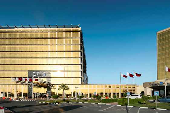 En moderne bygning med guldfarvede glasvægge, omgivet af flagstænger og en parkeringsplads. (AI-genereret tekst)