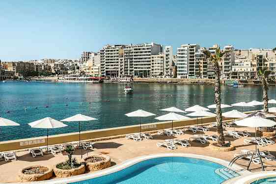 En udsigt over en pool med parasoller og liggestole, med en havn og by i baggrunden. (AI-genereret tekst)