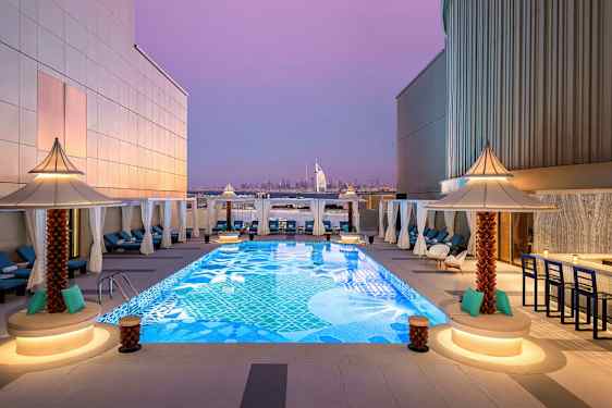 En tagterrasse med en dekorativ pool omgivet af liggestole og små parasoller. I baggrunden kan Burj Al Arab og Dubais skyline ses under en lilla aftenshimmel. (AI-genereret tekst)