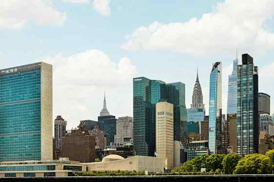 Billedet viser en bysilhuet med flere høje bygninger og skyskrabere omgivet af grønne områder under en klar himmel. (AI-genereret tekst)