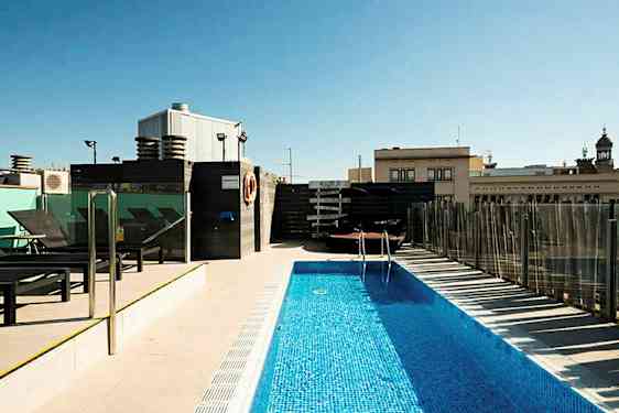 En udendørs pool på en tagterrasse omgivet af liggestole og bygninger i baggrunden. (AI-genereret tekst)