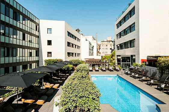 En hotelpool omgivet af liggestole og parasoller, med moderne bygninger omkring. (AI-genereret tekst)