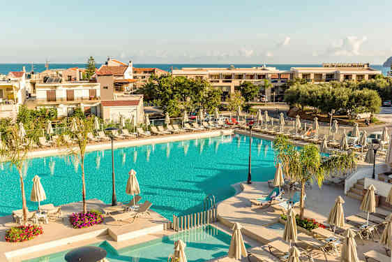 En udendørs swimmingpool omgivet af solsenge og parasoller med bygninger og havet i baggrunden. (AI-genereret tekst)