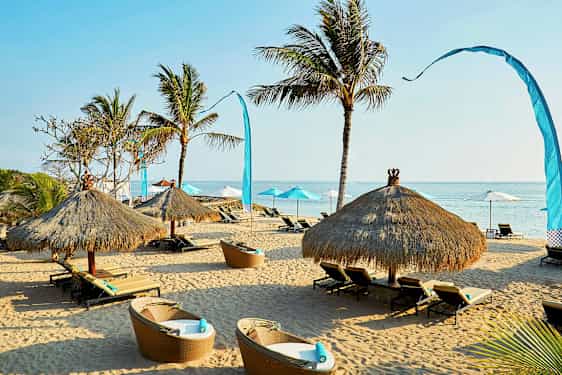 En sandstrand med liggestole og stråtækte parasoller omgivet af nogle palmer. (AI-genereret tekst)