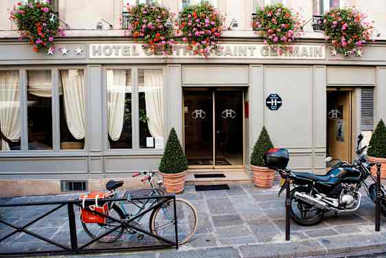 Eksteriøret af et hotel med skiltet 'Hotel Central Saint Germain' med blomsterkrukker over indgangen samt en cykel og motorcykel parkeret foran. (AI-genereret tekst)