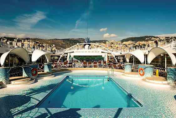 En pool på et krydstogtskib med solstole omkring og en by i baggrunden. (AI-genereret tekst)