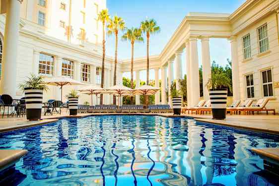 Poolområde med glinsende blåt vand, liggestole og parasoller omgivet af søjler og palmer. (AI-genereret tekst)