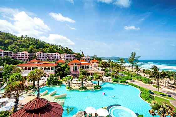 Billede af et luksuriøst hotelområde ved havet med en stor pool omgivet af palmer og bygninger med røde tage. (AI-genereret tekst)