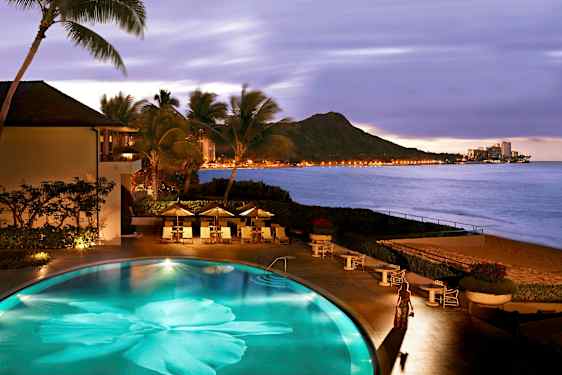 En pool ved stranden omgivet af palmer, med udsigt over bjerge og lysene fra en by ved havet. (AI-genereret tekst)