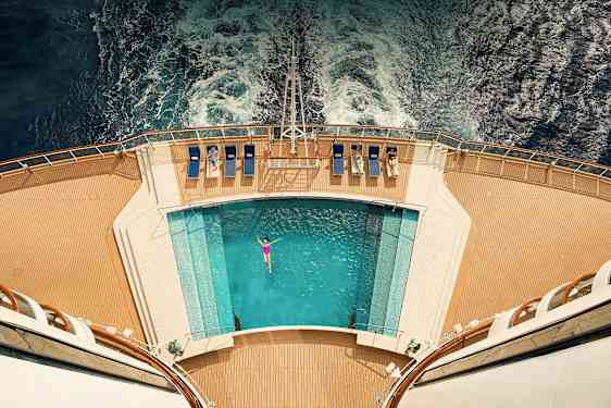 Udsigt oppefra af et skibsdæk med pool og liggestole, vand bagved. (AI-genereret tekst)