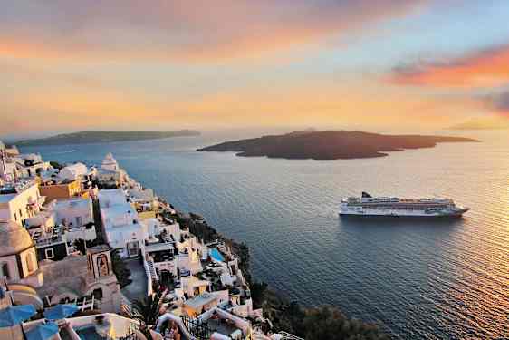 Hvidkalkede bygninger på en klippe over havet med et krydstogtskib i baggrunden under en farverig solnedgang. (AI-genereret tekst)