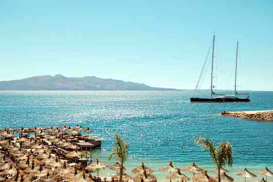 Strand med parasoller og lang sejlbåd på havet med bjerge i baggrunden. (AI-genereret tekst)