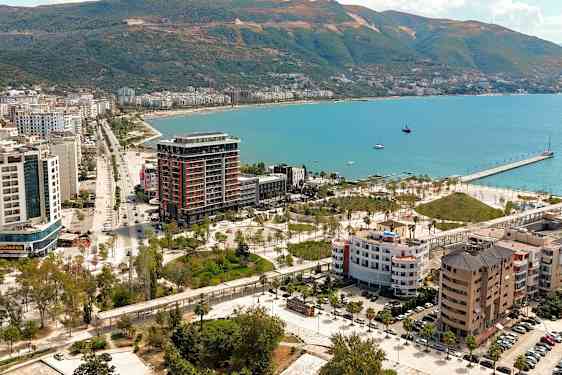 Udsigt over en kystby med bygninger, grønne områder og en kaj ved havet. (AI-genereret tekst)