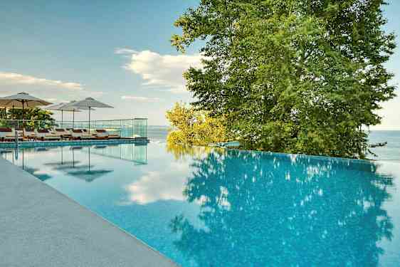 En infinitypool med liggestole og parasoller ved siden af, omgivet af træer og med udsigt over havet. (AI-genereret tekst)