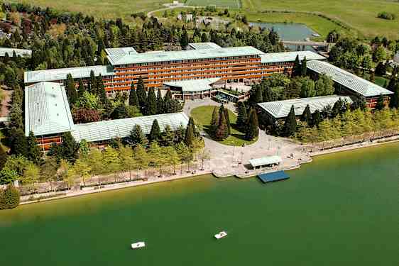 Luftfoto af en stor bygning omgivet af grønne træer ved en vandoverflade med små både. (AI-genereret tekst)