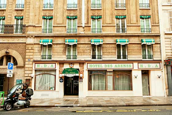 Bygning med hotellfacade, skilt med teksten 'Hotel des Arenes' og grønne og hvide markiser over vinduerne. (AI-genereret tekst)