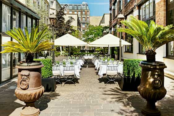 Udendørs selskabsområde med borde og parasoller omgivet af bygninger og palmer. (AI-genereret tekst)