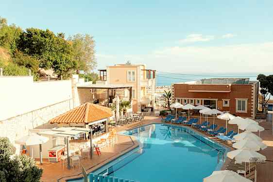 En hotelpool med liggestole og parasoller ved siden af samt bygninger og vegetation i baggrunden. (AI-genereret tekst)