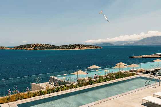 Billede af en infinitypool med udsigt over havet og omkringliggende øer. Et glasrækværk omgiver poolområdet, og parasoller er placeret ved poolkanten. (AI-genereret tekst)