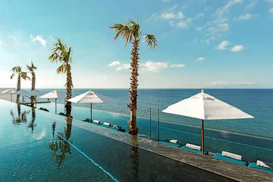 En infinitypool med udsigt over havet, omgivet af parasoller og palmer under en klar blå himmel. (AI-genereret tekst)