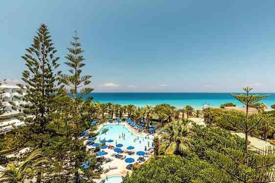 Billede af hotelområde med pool og solsenge omgivet af grønne områder og udsigt over havet. (AI-genereret tekst)