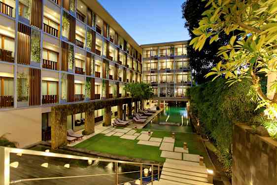 Et hotelområde om aftenen med oplyste vinduer, en rektangulær pool omgivet af grønne planter og liggestole samt en velholdt græsplæne. (AI-genereret tekst)