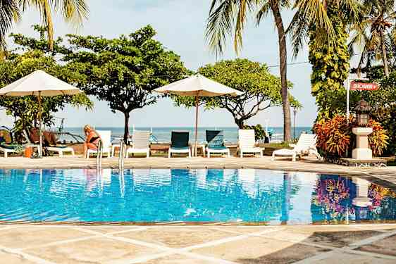 Billede af en pool med klart blåt vand, omgivet af liggestole og parasoller med havet og palmetræer i baggrunden. (AI-genereret tekst)