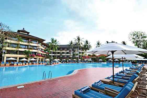 Billede af en udendørs pool omgivet af liggestole og parasoller med et hotel i flere etager og palmer i baggrunden. (AI-genereret tekst)