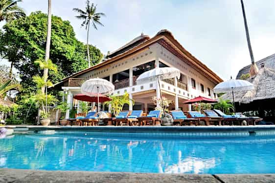 En stor villa med stråtag og en pool foran, omgivet af liggestole og parasoller. (AI-genereret tekst)