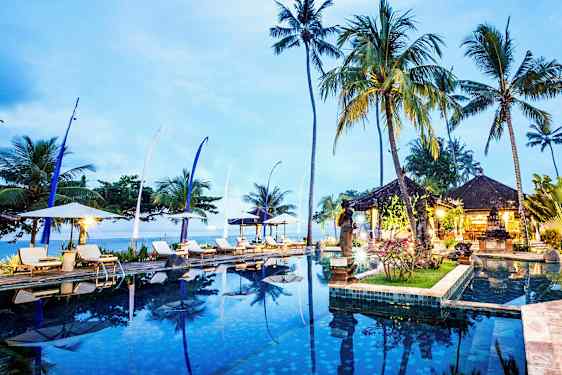 En infinity-pool omringet af liggestole, parasoller, høje palmer og et åbent pavillonområde ved skumring. (AI-genereret tekst)