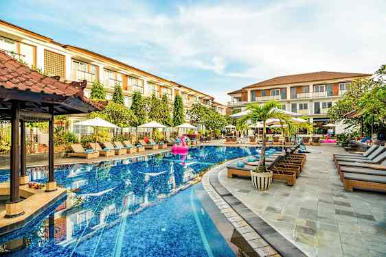 En hotelpool med liggestole og parasoller omkring, samt nogle bygninger i baggrunden. (AI-genereret tekst)