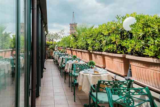 En udendørs terrasse med dækkede borde og grønne metalstole, kantet af store plantekasser med grønne planter. (AI-genereret tekst)