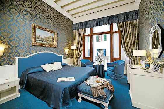 Et smukt indrettet hotelværelse med blå og gyldne detaljer, stor seng, siddeområde og store vinduer. (AI-genereret tekst)