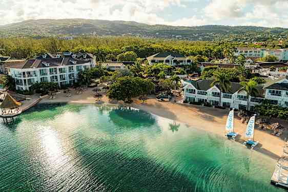 Flere hotelbygninger ved en rolig sandstrand med klart vand og udsigt over frodige bakker. (AI-genereret tekst)