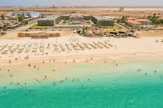 Luftfoto af en strand med turkis vand, liggestole og parasoller, samt bagved en hotelbygning. (AI-genereret tekst)