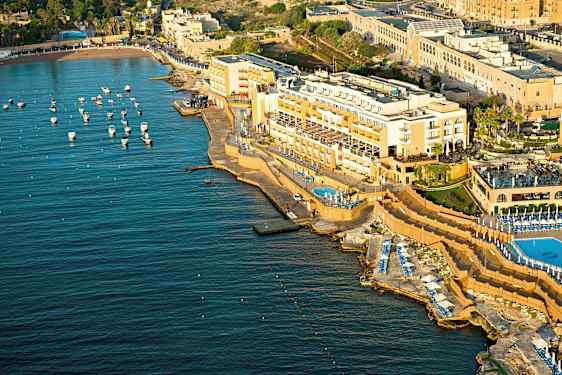Luftfoto af et kysthotel med pools og liggestole ved havet. Små både er fortøjet i vandet. (AI-genereret tekst)
