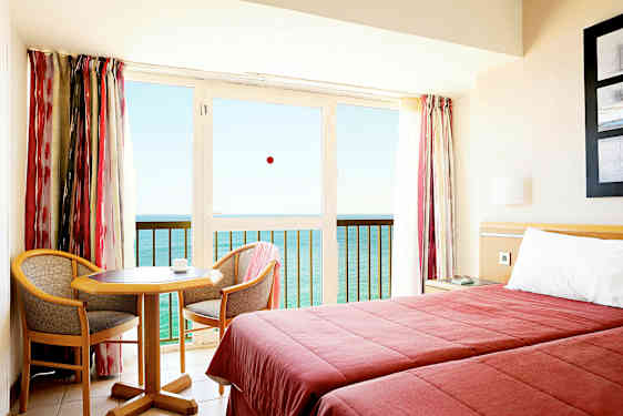 Et hotelværelse med to senge, et lille bord og to stole, samt en balkon med udsigt over havet. (AI-genereret tekst)