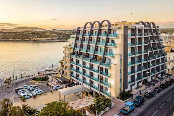 En bygning ved vandet med moderne etager og altaner. Foran bygningen ses parkerede biler og en vej. (AI-genereret tekst)