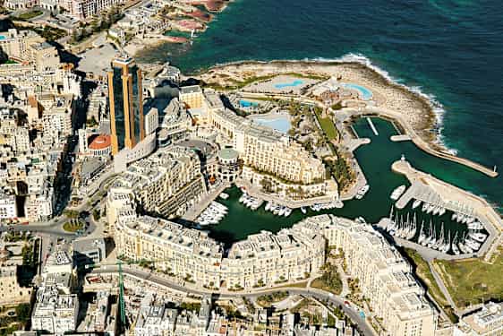 Luftfoto af en kystby med høje bygninger, småbådehavn og havet i baggrunden. (AI-genereret tekst)