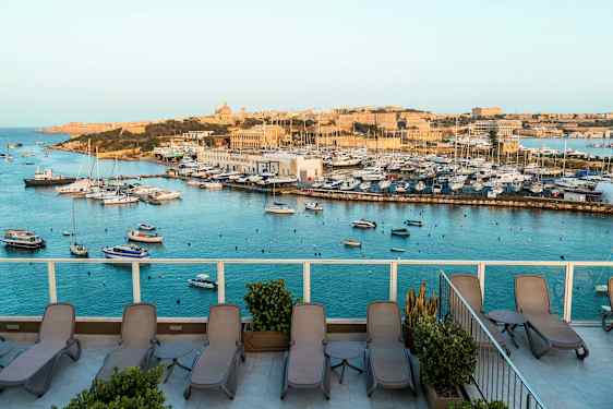 Udsigt over en havn med både og en terrasse med liggestole. (AI-genereret tekst)