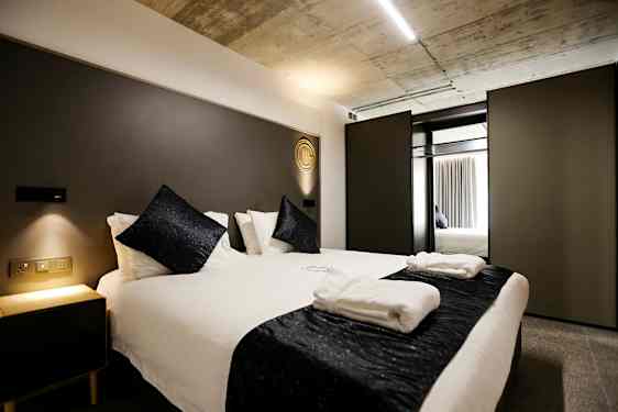 Soveværelse med en moderne dobbeltseng, hvidt sengetøj og mørke dekorative puder. Et natbord med blød belysning står ved siden af. (AI-genereret tekst)