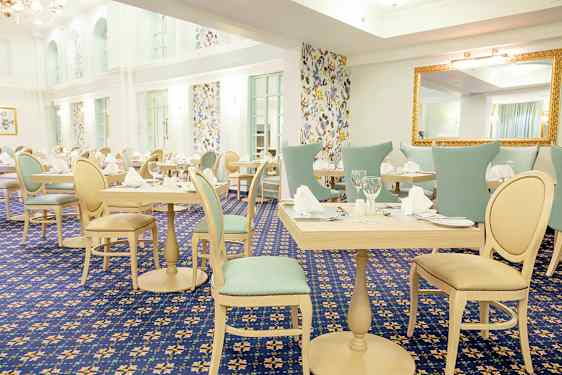 Et lyst og elegant indrettet spisesal med dækkede borde og stole, omgivet af vægge med mønstrede tapeter. (AI-genereret tekst)