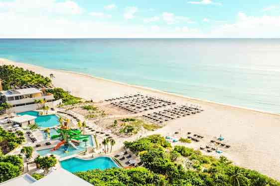 Billede af en sandstrand ved havet med blåt vand. På stranden er der liggestole og parasoller. I forgrunden ses en pool med vandrutsjebaner samt omgivelser med grønne planter. (AI-genereret tekst)