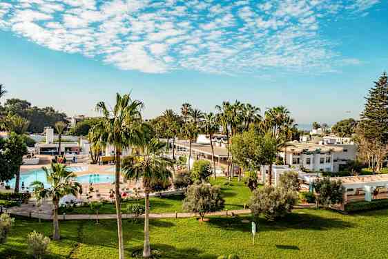 Hotelområde med græsplæner, palmer, pool og hvide bygninger. (AI-genereret tekst)